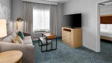 A hotel room with a sofa, a small table, a TV on a wooden stand, a bed, and a window with curtains.