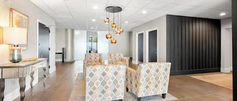 A spacious hotel lobby with a modern design, featuring a seating area with patterned chairs, a central light fixture, and a large mirror.