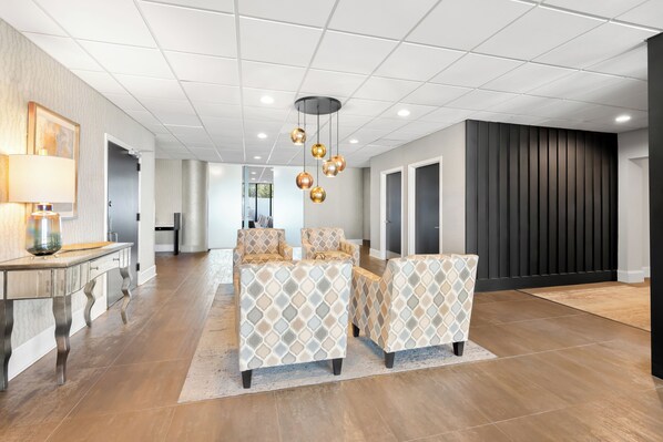 A spacious hotel lobby with a modern design, featuring a seating area with patterned chairs, a central light fixture, and a large mirror.