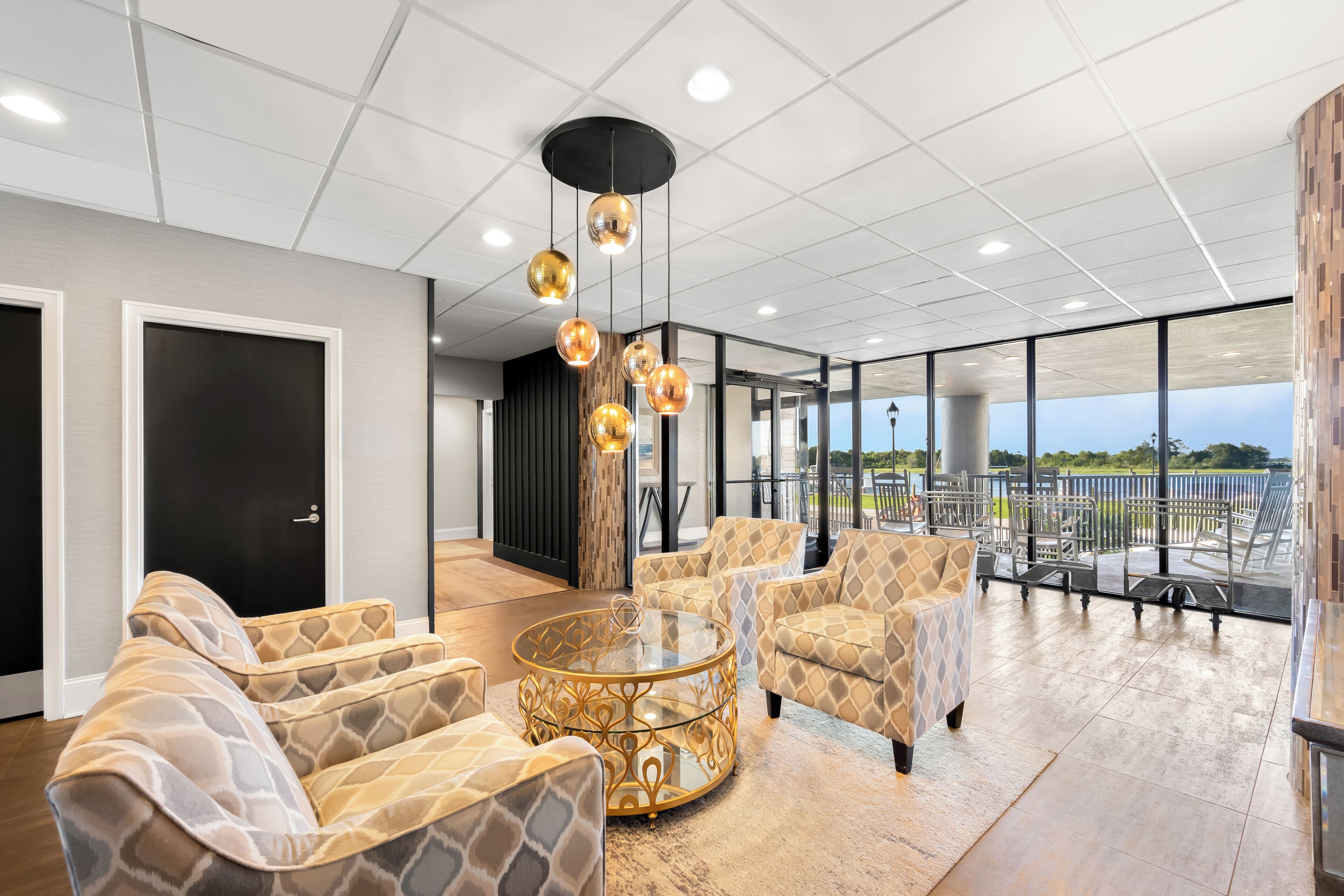 A modern interior with patterned furniture, a glass coffee table, pendant lighting, and a view through large windows.
