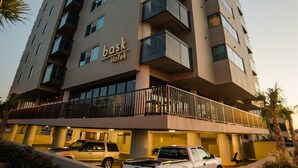 Un immeuble hôtelier à plusieurs étages, doté de balcons, d’un parking et d’une enseigne indiquant « Bask Hotel ».