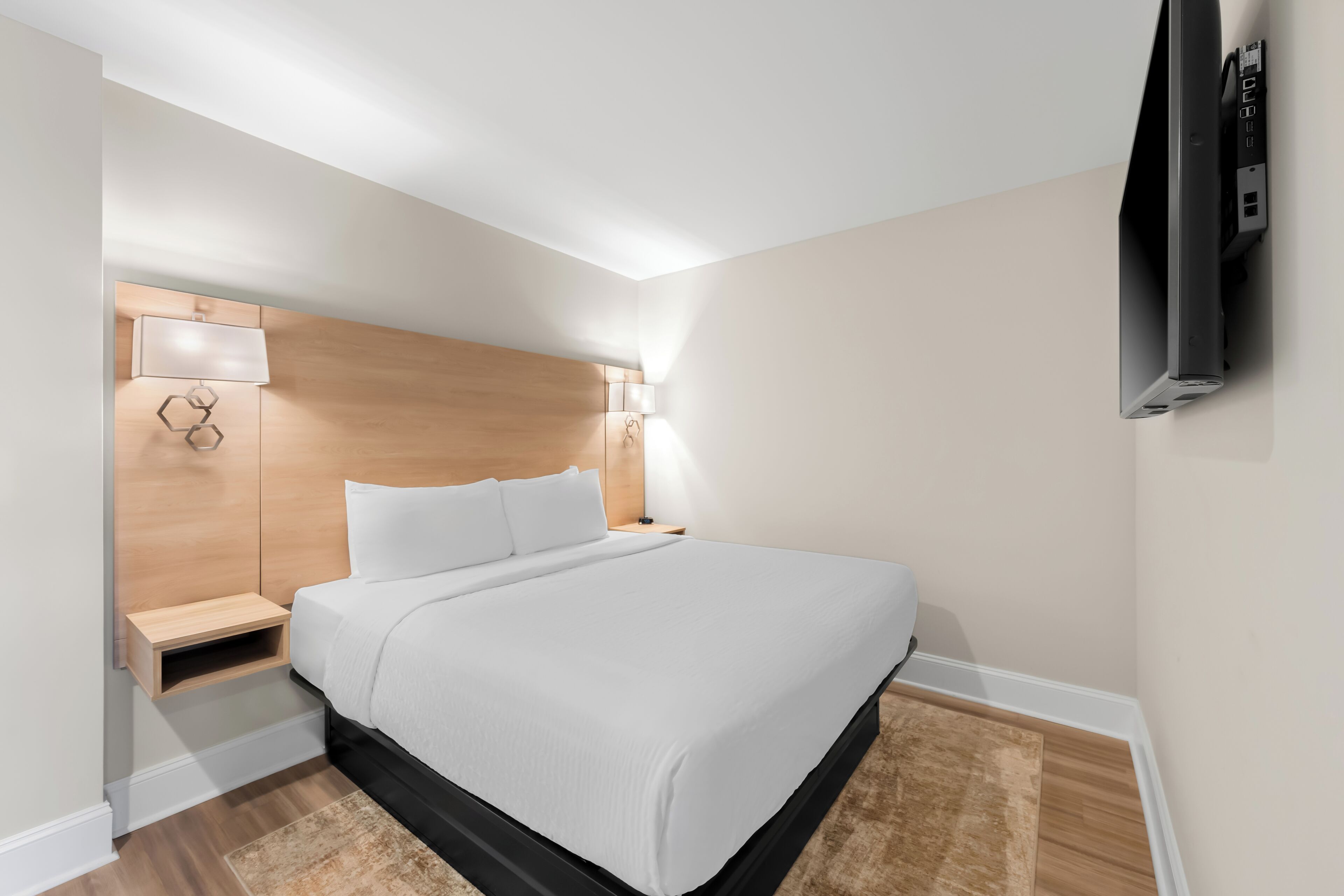 A modern hotel room with a bed, bedside tables, a TV, and a wall-mounted lamp.