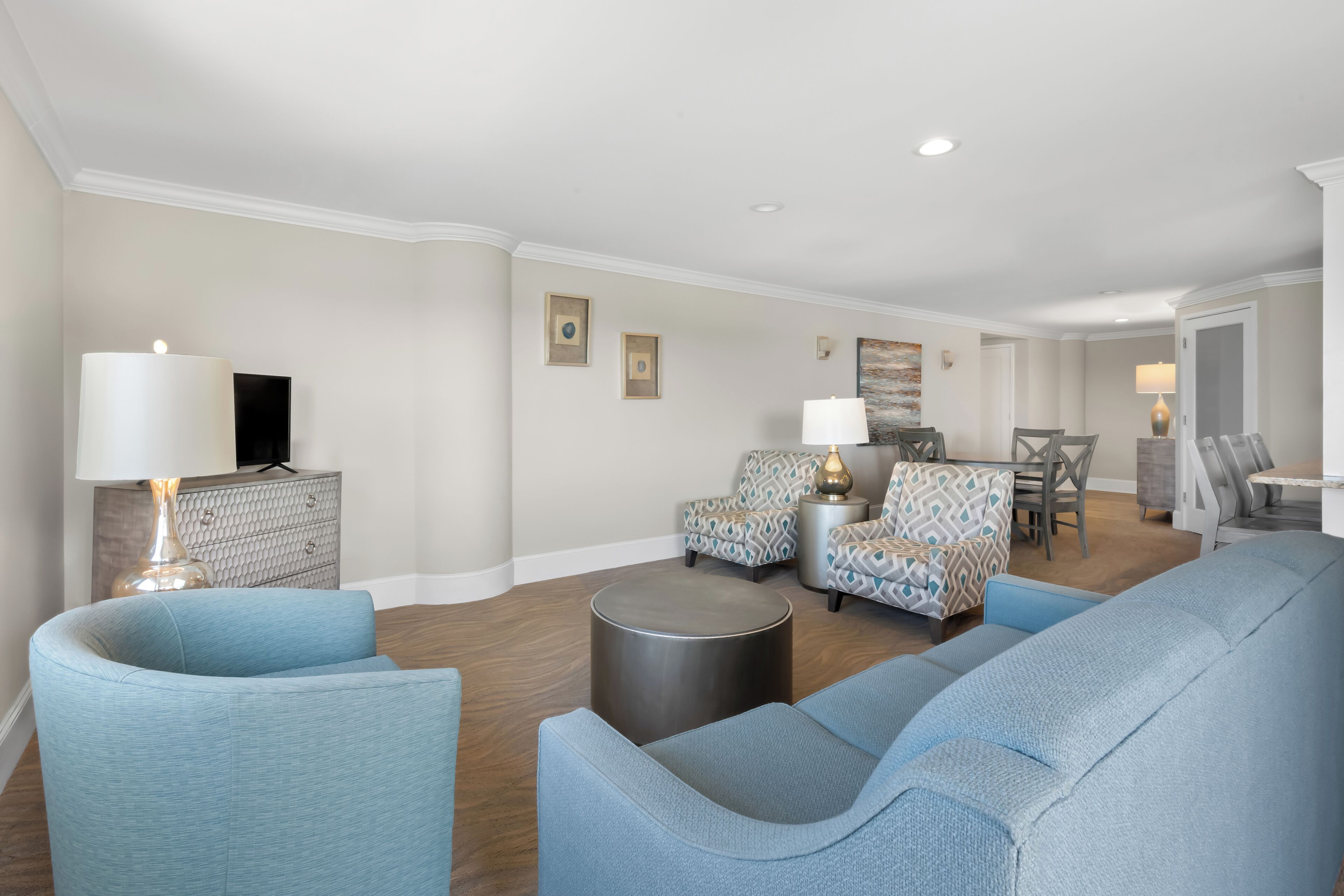 A modern living room with a flat-screen TV, two patterned armchairs, a sofa, a coffee table, and a dining area with a table and chairs.