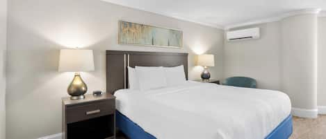 Una habitación de hotel con una cama, mesitas de noche, lámparas, un cuadro y un equipo de aire acondicionado.