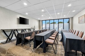 Un salón de conferencias con una larga mesa, sillas, un televisor de pantalla plana y ventanas amplias.