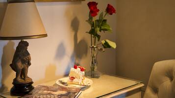 A table with a book, a plate with cake, a vase with roses, a lamp, and a painting on the wall.