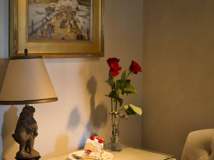 A table with a book, a plate with cake, a vase with roses, a lamp, and a painting on the wall.