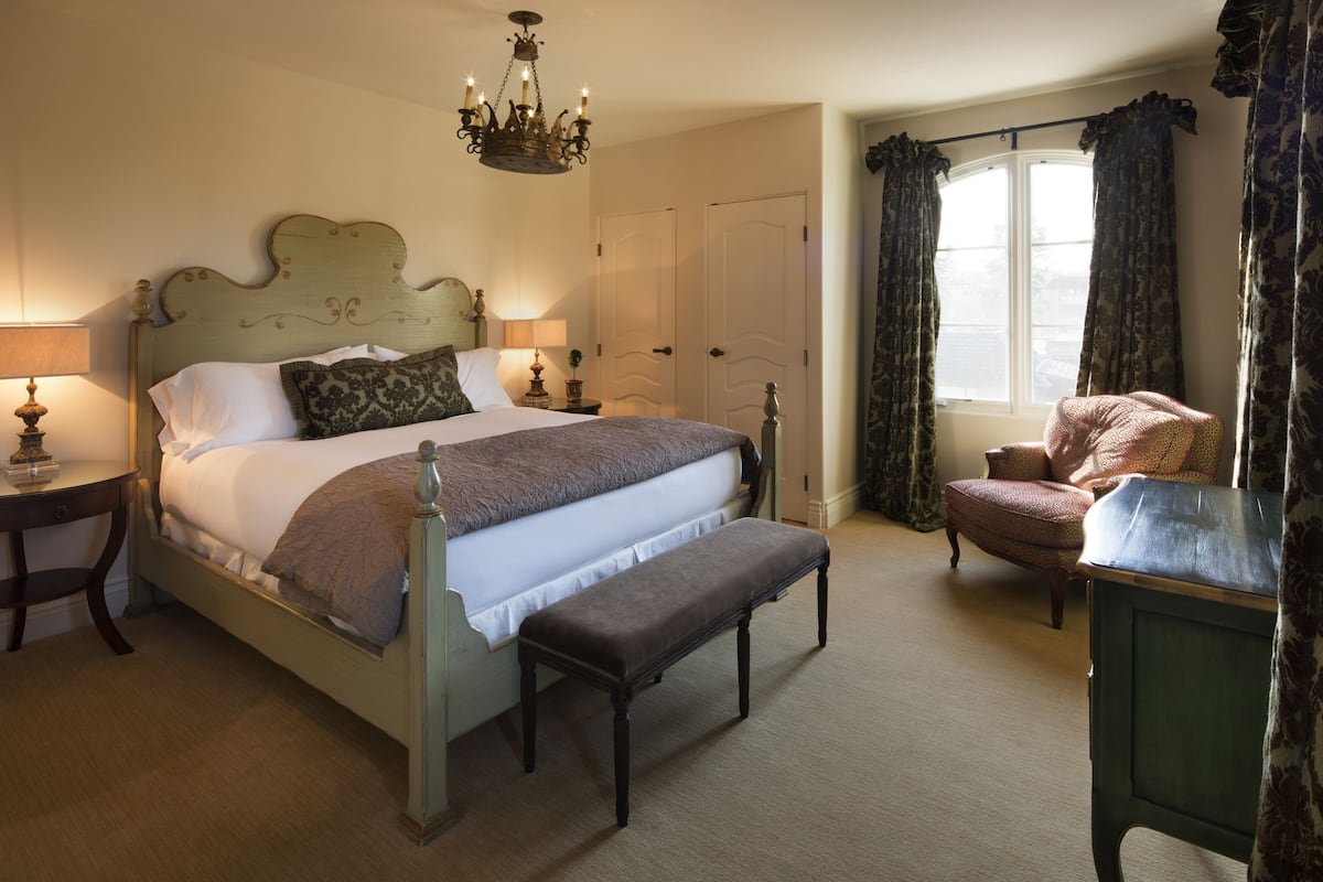 A bedroom with a four-poster bed, a bench, a chair, a table, and a window with curtains.