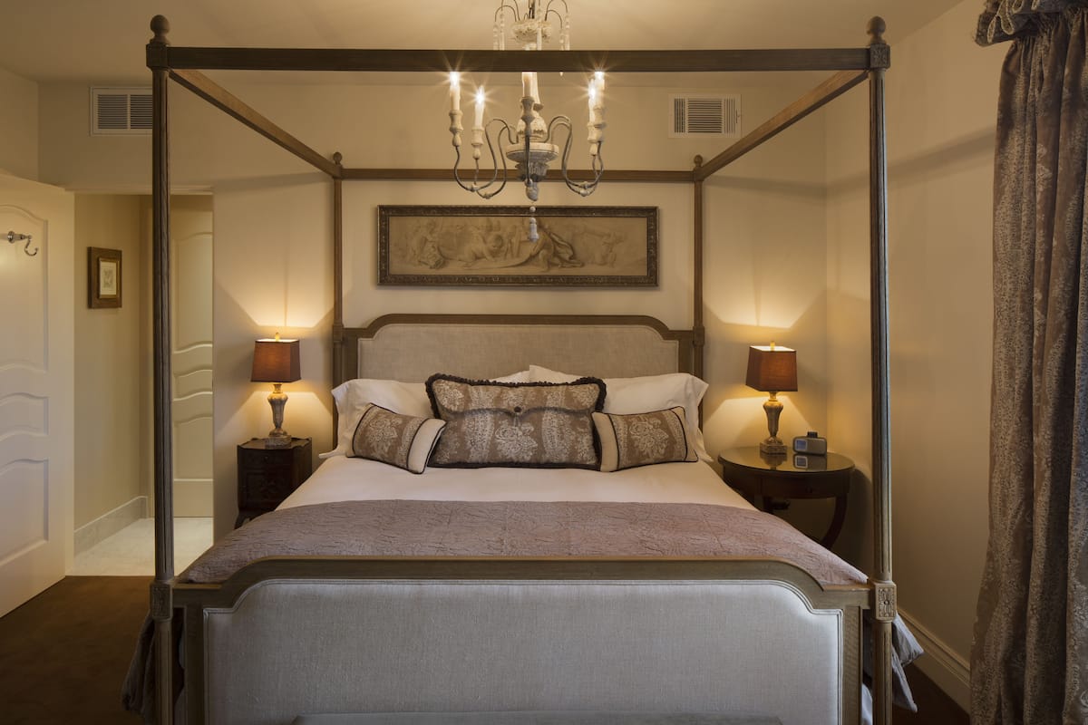 A four-poster bed with a chandelier, two bedside tables with lamps, a framed artwork on the wall, and a door with a coat hook.