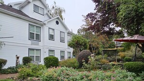 Una casa blanca de dos pisos con un jardín cuidado, que incluye una variedad de plantas florecientes y un pequeño pabellón.
