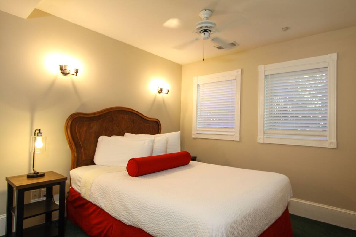 A bedroom with a wooden headboard, white bedding, a red pillow, two wall-mounted lights, and a ceiling fan.