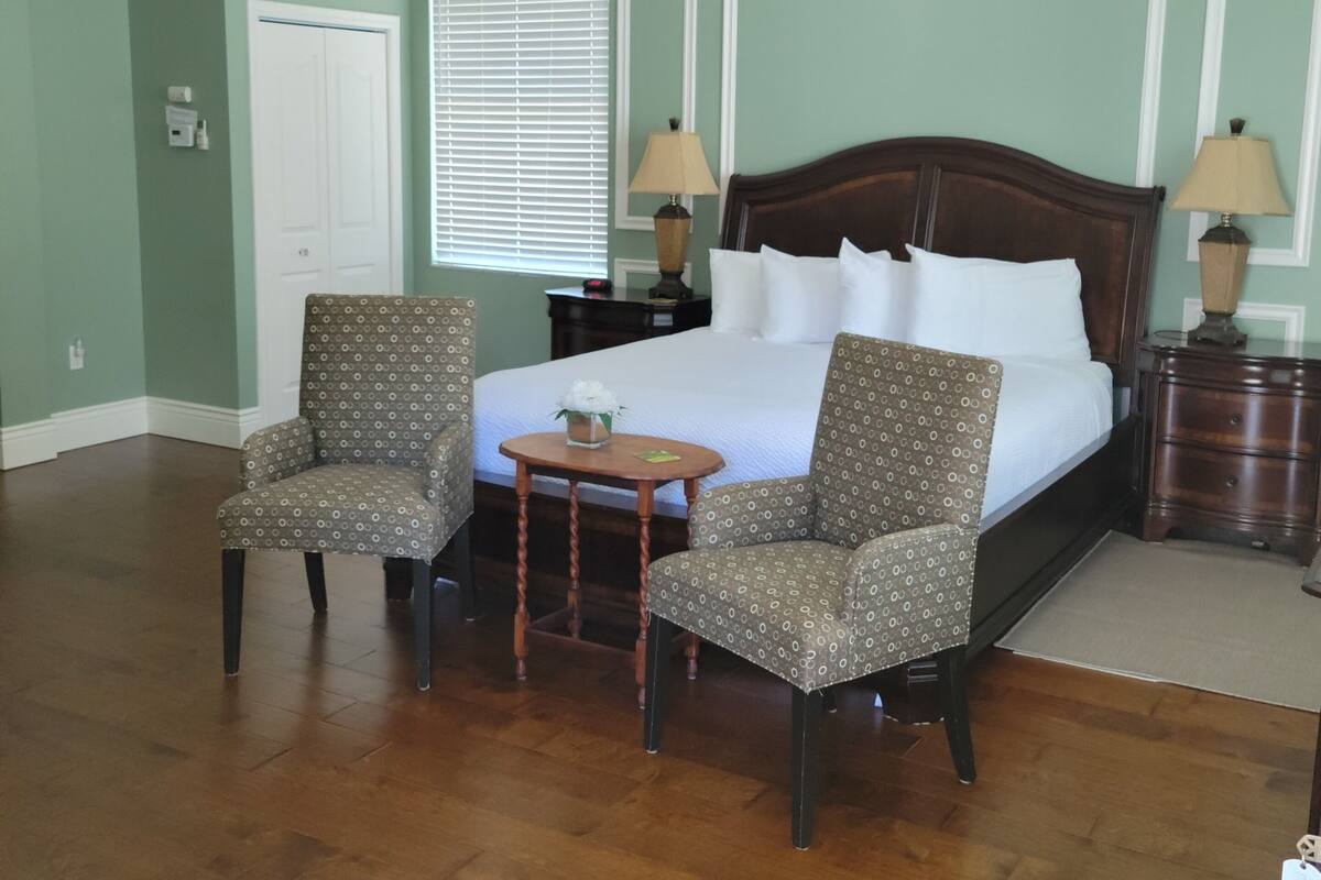 A hotel room with a large bed, two armchairs, a small table, a ceiling fan, and a window with blinds.