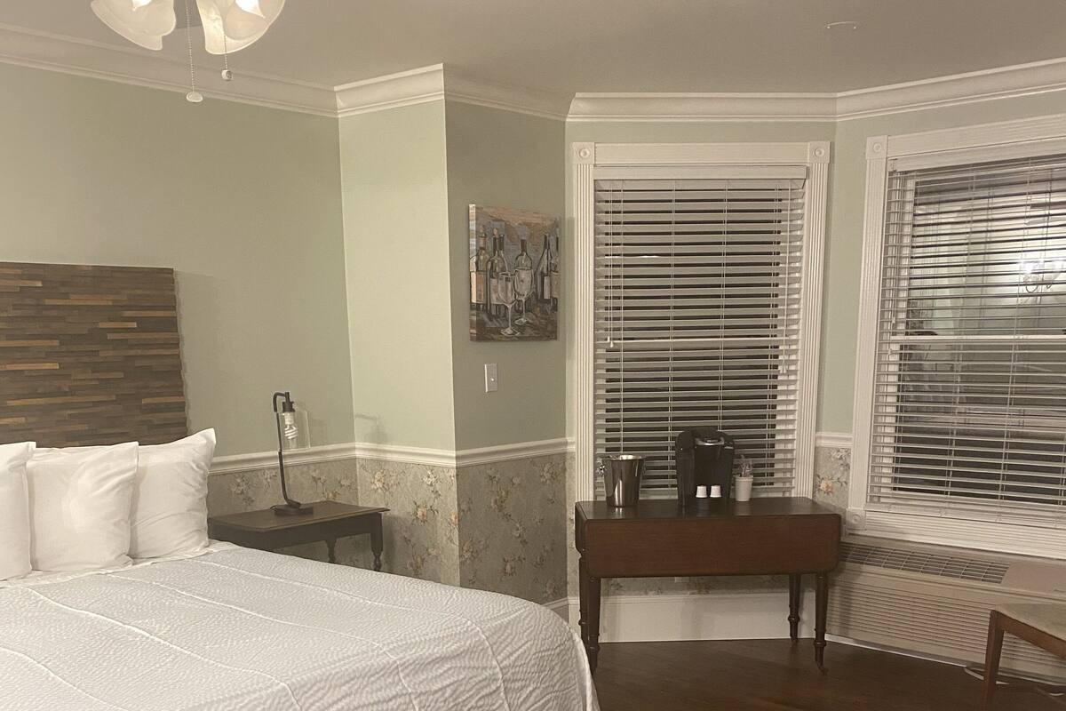 A bedroom with a large bed, a wooden floor, a ceiling fan, a nightstand, a bench, and a window with blinds.