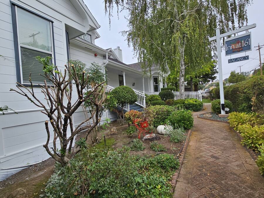 A two-story house with a front porch, a garden with various plants and a small tree, and a sign indicating a vacancy.