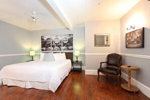 A hotel room with a large bed, two bedside tables with lamps, a chair, a small round table, a ceiling fan, and artwork on the walls.