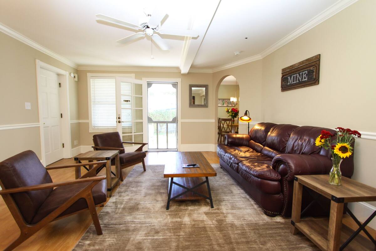A living room with a brown leather sofa, wooden coffee table, and matching chairs. There is a ceiling fan, a vase with flowers, and a sign on the wall.