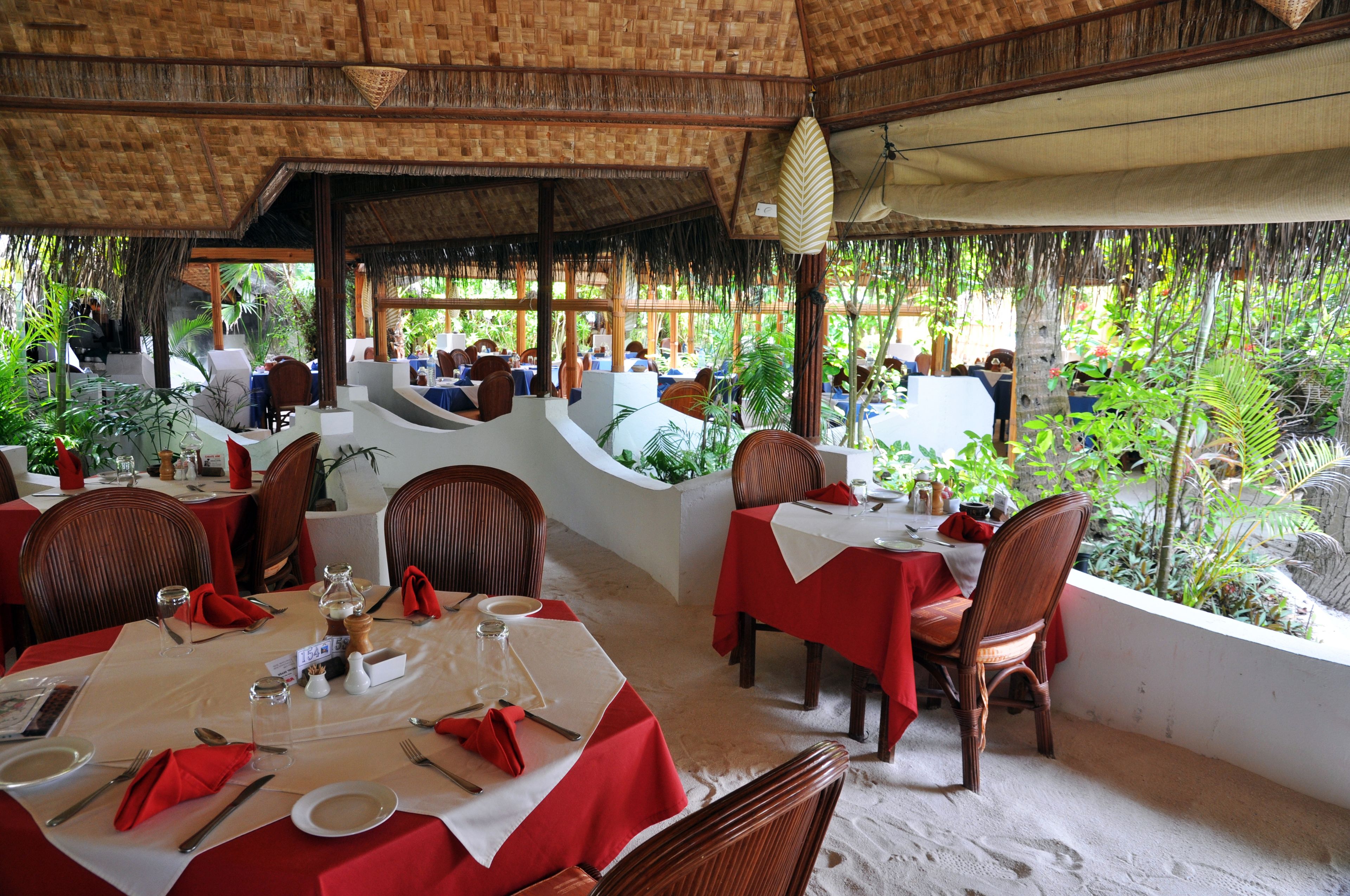 A restaurant with tables set for dining, surrounded by lush greenery and a thatched roof.