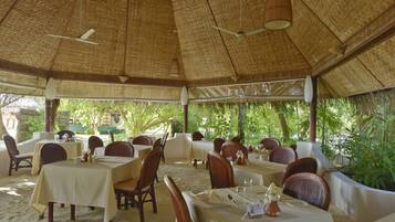 An outdoor dining area with tables and chairs, a thatched roof, and greenery in the background.