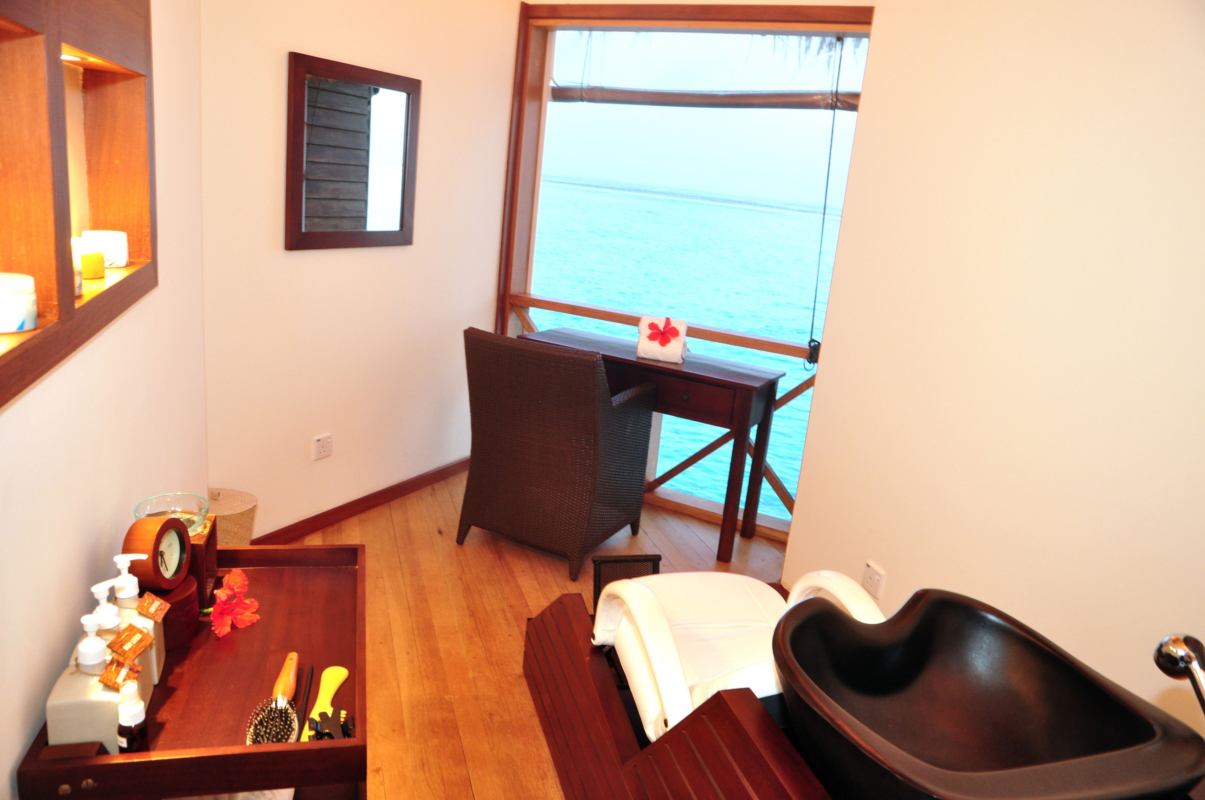 A room with a wooden desk, a chair, a small table with toiletries, a large window with a view of the sea, and a small black bathtub.