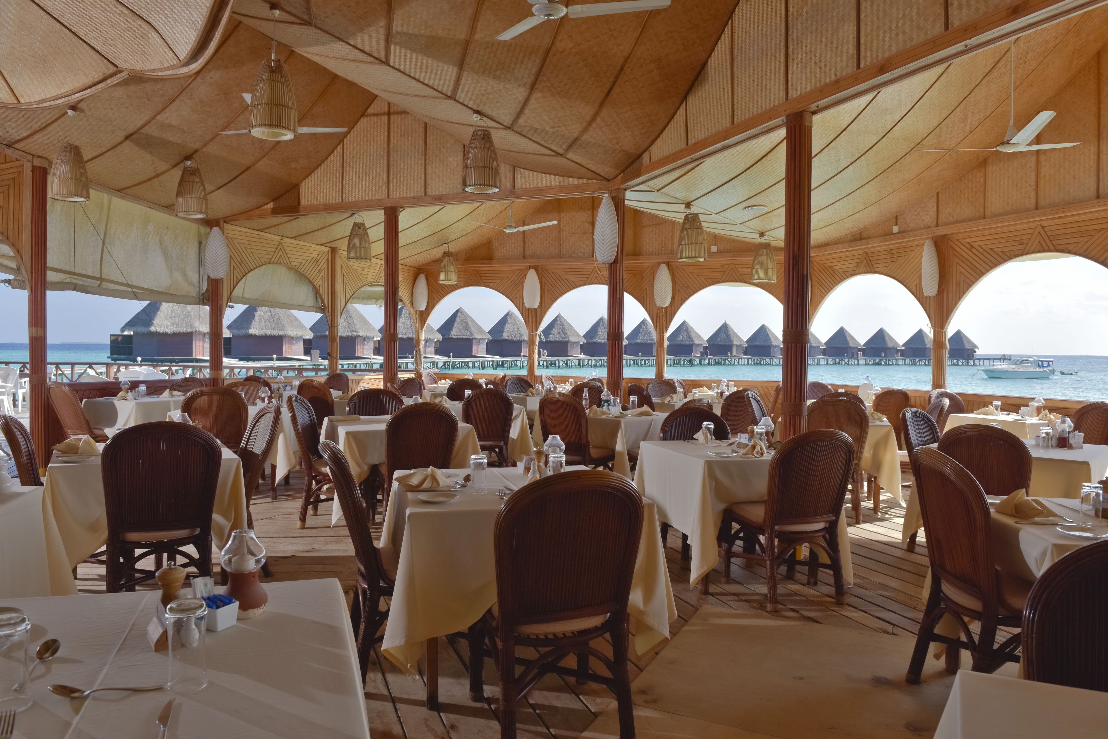 An indoor dining area with tables and chairs, large windows, and a view of overwater bungalows.
