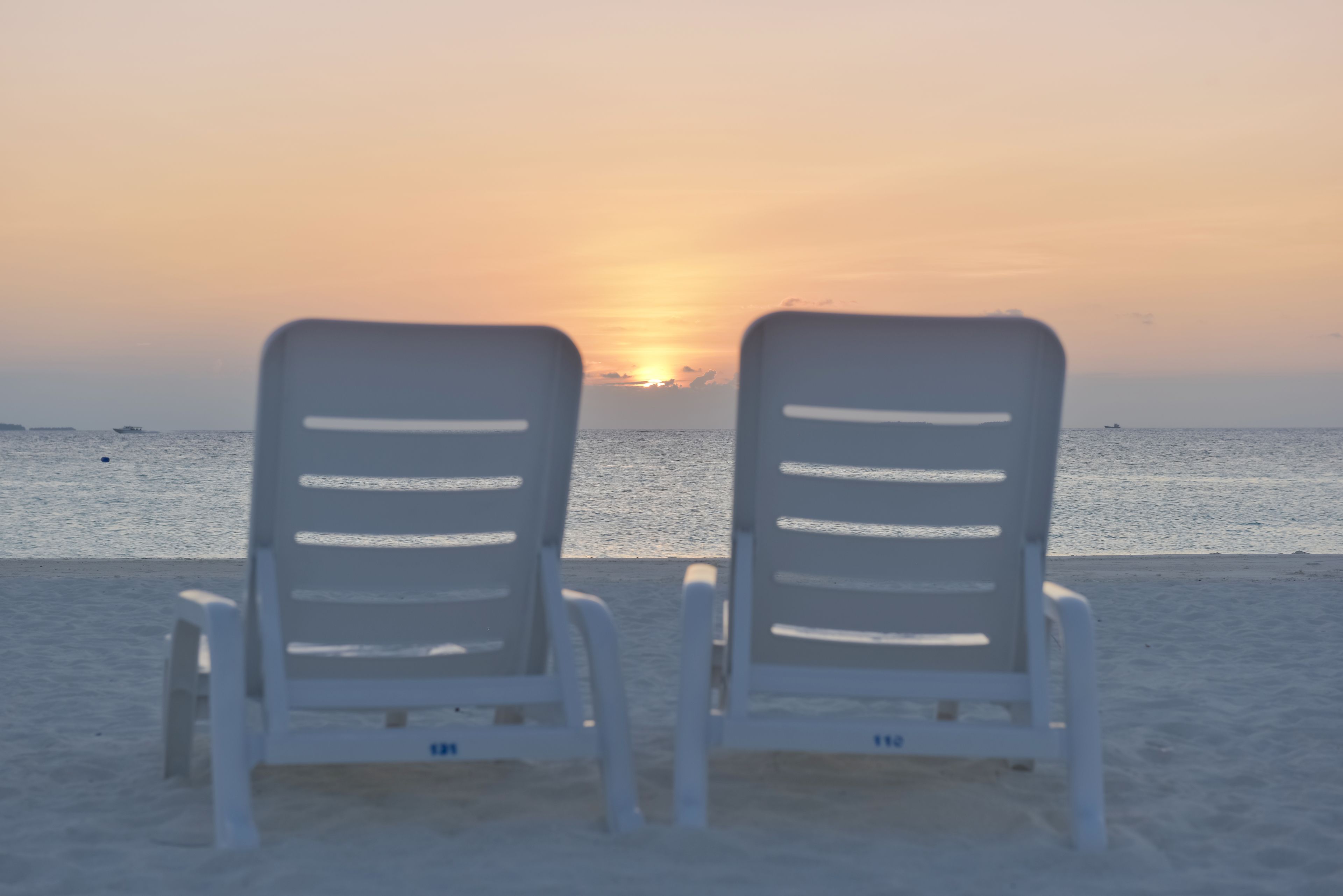 Zwei weiße Liegestühle, die auf einen Sonnenuntergang über dem Meer zeigen.