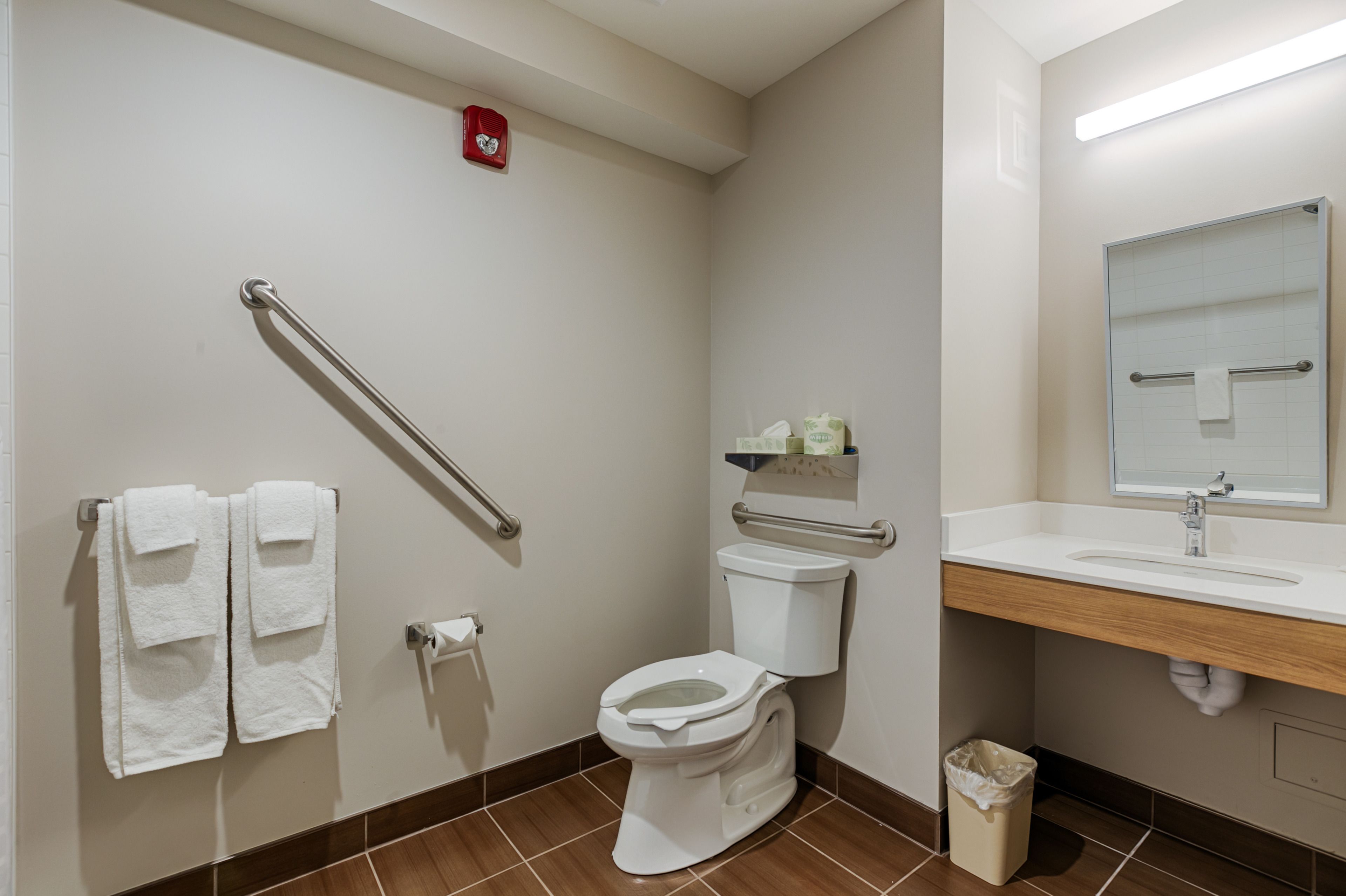 Une salle de bain équipée d’un WC, d’un lavabo, d’un miroir, d’une barre d’appui et d’une poubelle.