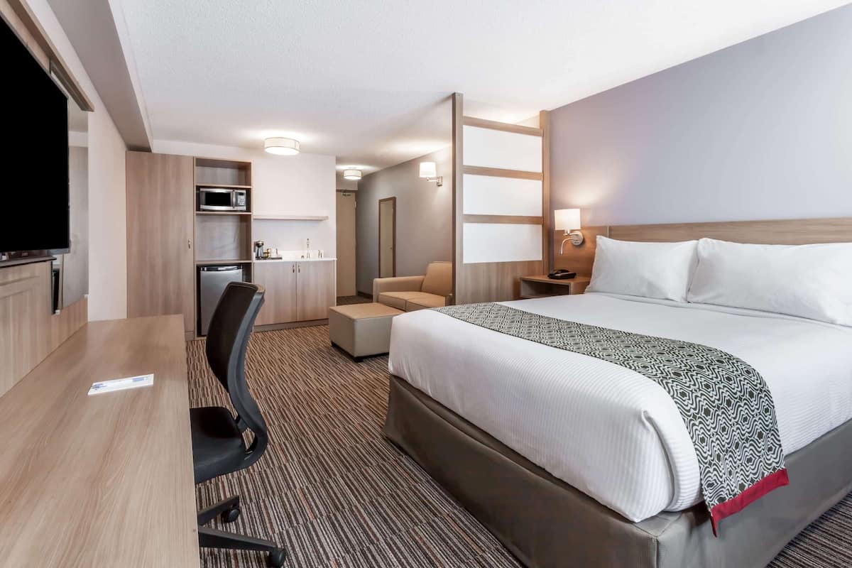 A hotel room with a bed, a desk, a chair, a microwave, a small kitchenette, and a TV.