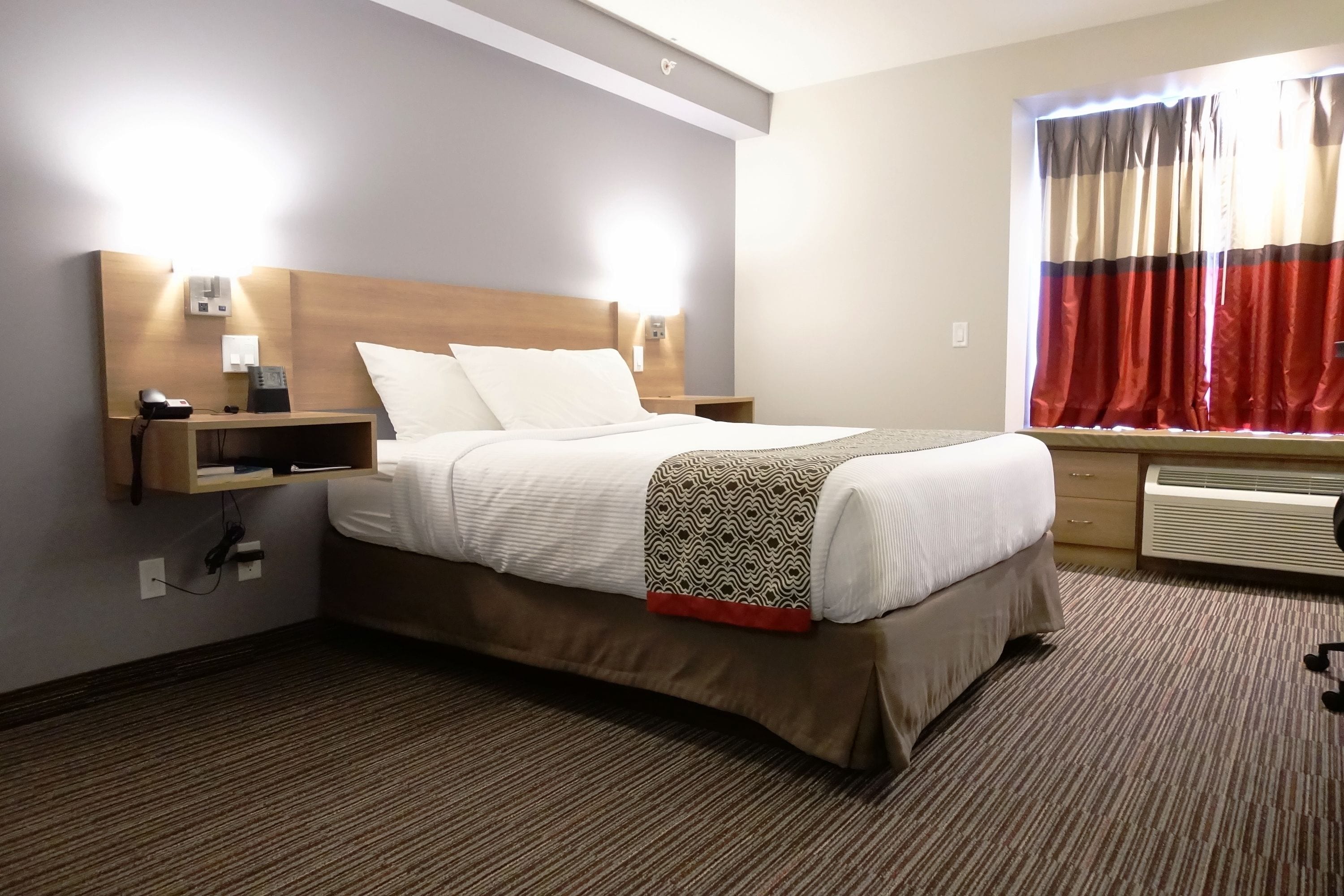 A hotel room with a bed, bedside tables, a wall-mounted heater, and a window with curtains.