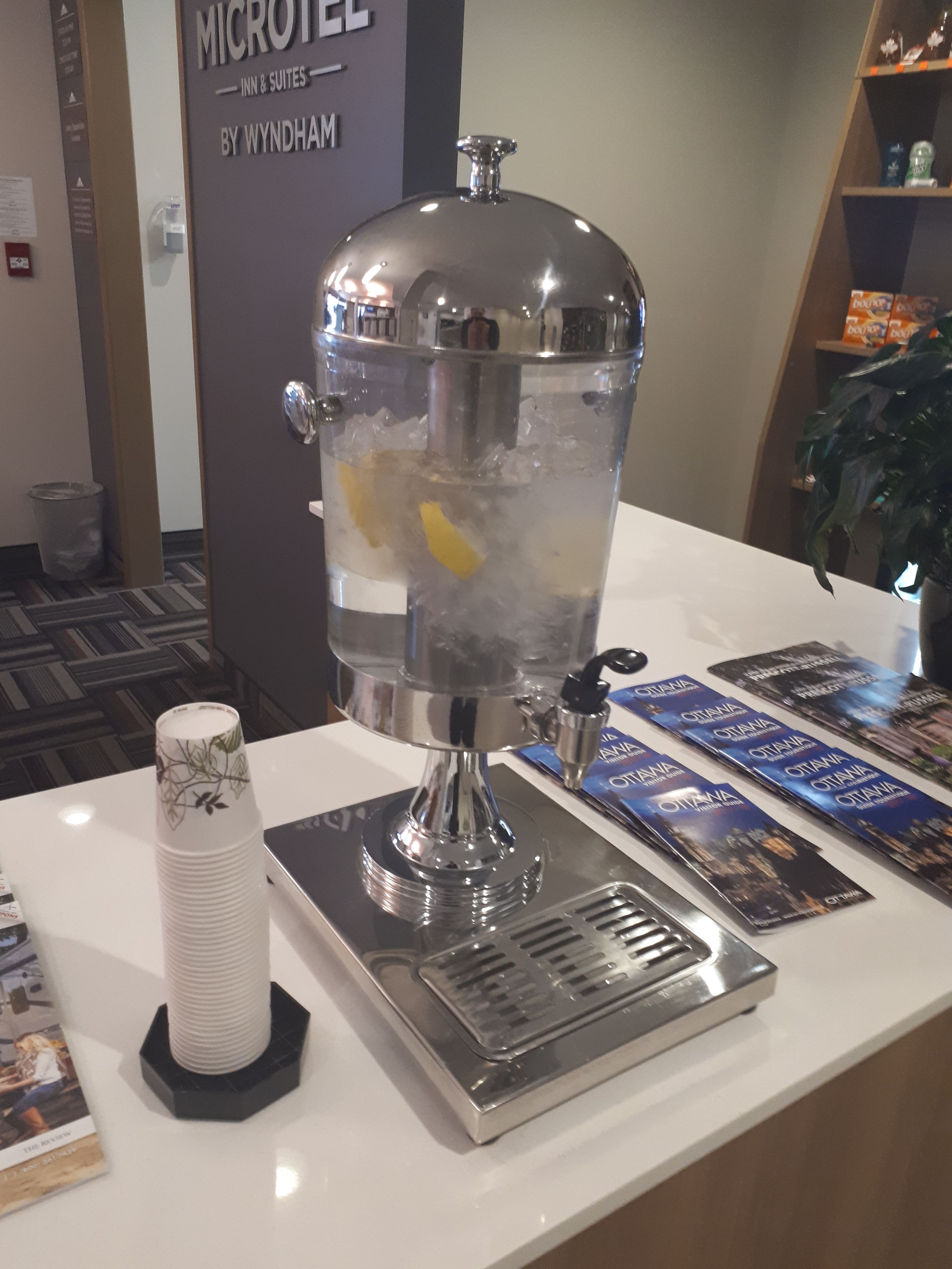 Een minibarschaal met een groot ijsdispenser, een stapel witte bekers en brochures op een witte toonbank.