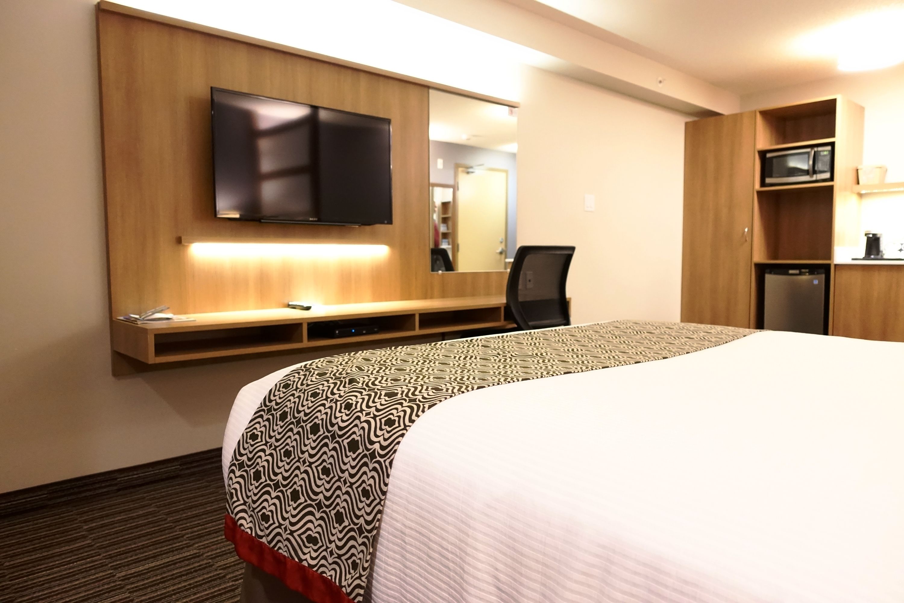 A hotel room with a large bed, a flat-screen TV mounted on the wall, a microwave, and a small kitchenette.