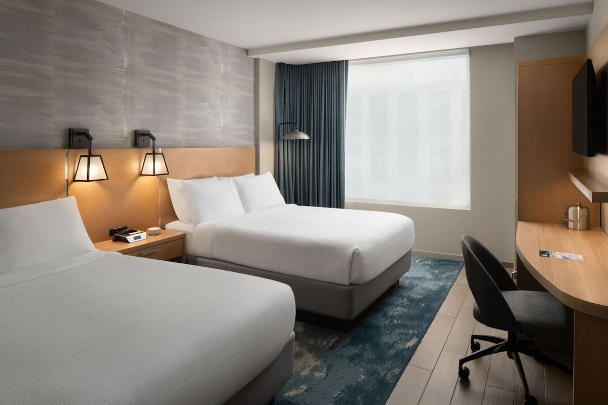 A hotel room with two beds, a desk, a chair, a nightstand with a lamp, and a large window with curtains.