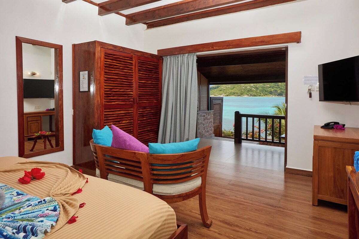 A bedroom with a wooden bed, a wooden chair with cushions, a TV, and a balcony with a view of the sea.
