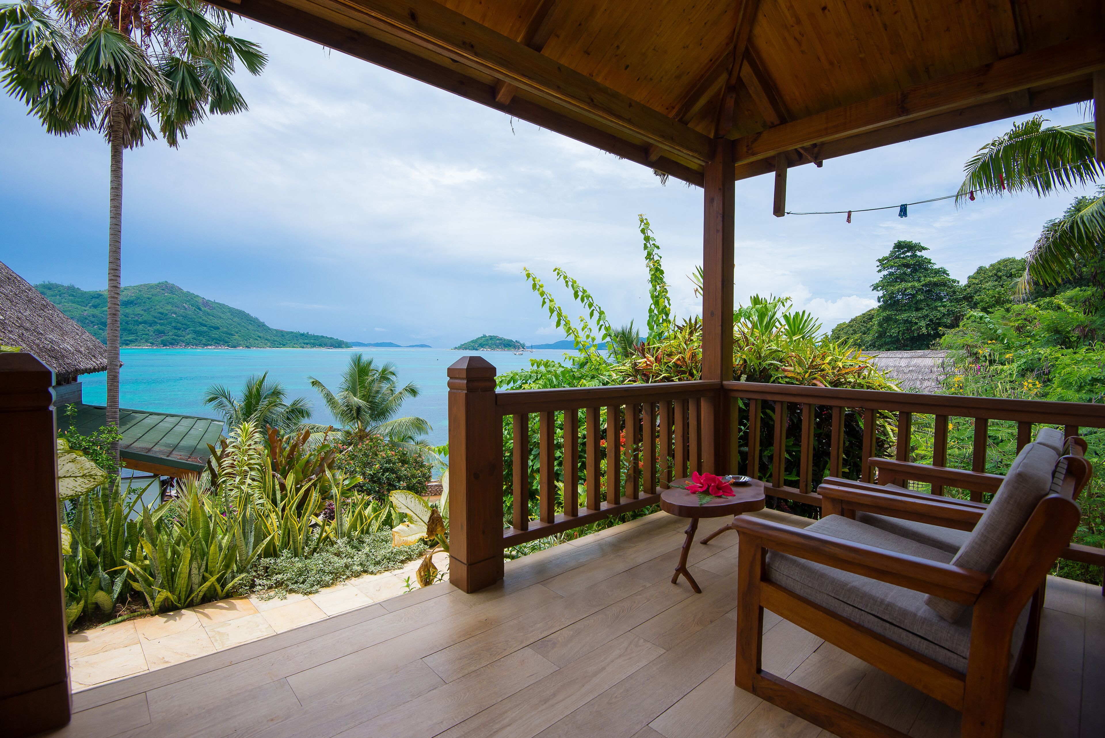 A wooden deck with a wooden bench and table, overlooking a beach with palm trees and a view of the ocean.