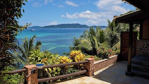 A view from a covered outdoor area with a stone wall and wooden railing, overlooking a clear blue sea and distant mountains. - Colibri Guest House (Praslin Island)