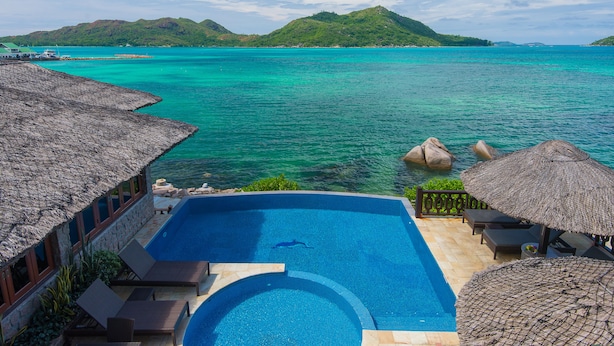 A poolside area with a clear view of the ocean and a mountainous landscape in the background. - Colibri Guest House (Praslin Island)