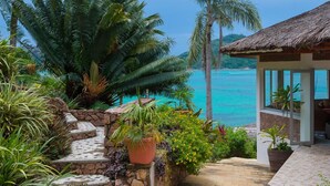 A tropical garden with a stone staircase leading to a thatched-roof building, overlooking a turquoise sea. - Colibri Guest House (Praslin Island)