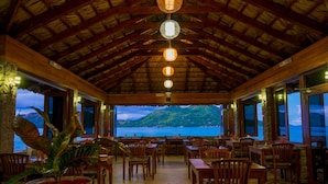 A restaurant with a thatched roof, wooden furniture, and a view of the sea. - Colibri Guest House (Praslin Island)