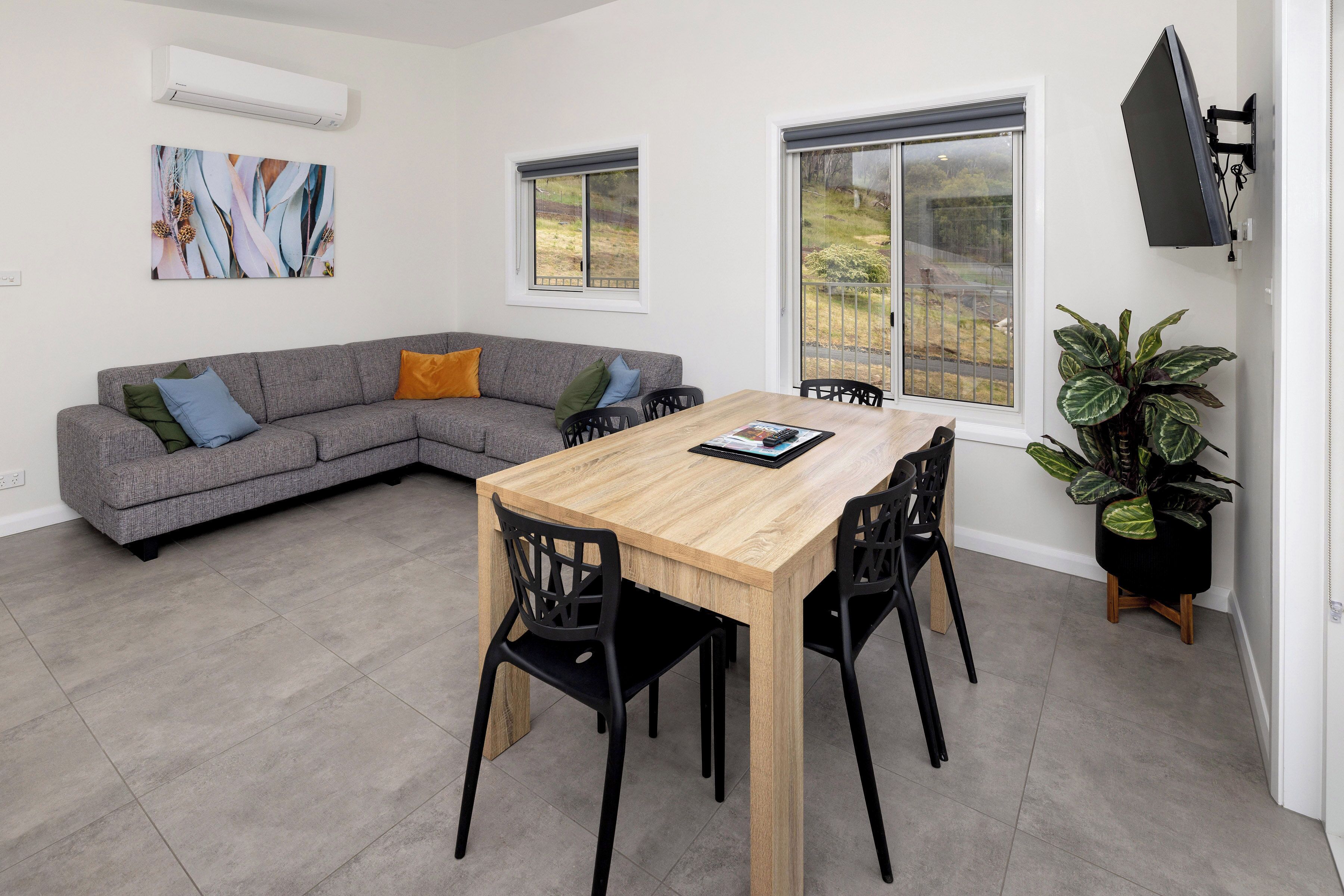 A modern living space with a wooden dining table, black chairs, a grey sectional sofa, and a large window offering a view of the outdoors.
