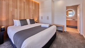 A modern bedroom with a large bed, wooden panel wall, built-in wardrobe, bedside table, and a bathroom with a mirror and sink. - BIG4 Launceston Holiday Park (South Launceston)