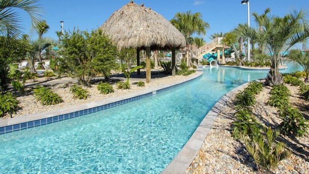 A clear blue pool with a thatched-roof structure, palm trees, and a water slide in the background. - ChampionsGate Platinum Resort Homes by Ocean Florida (Kissimmee)