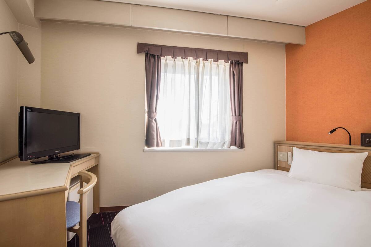 Una habitación de hotel con una cama, un televisor en un escritorio, una ventana con cortinas y una pared naranja.