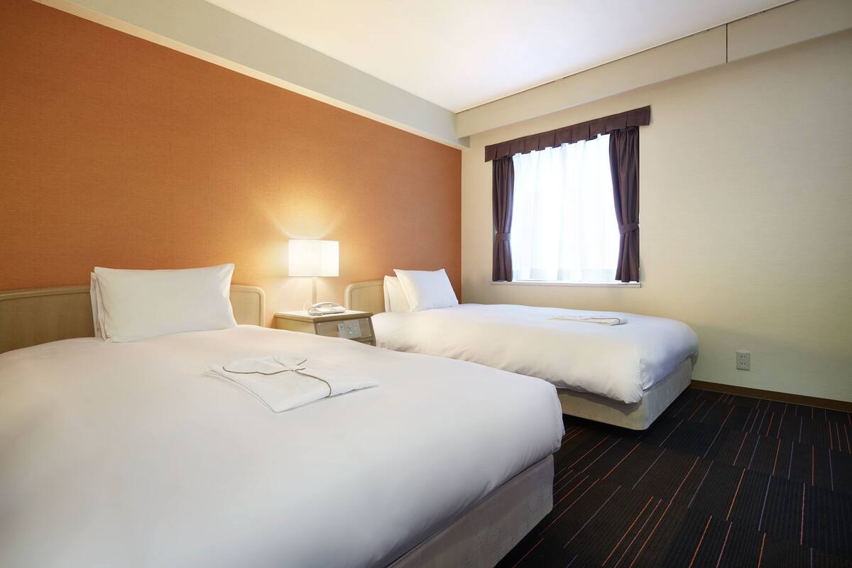 Una habitación de hotel con dos camas, una mesita de noche con lámpara y una ventana con cortinas.