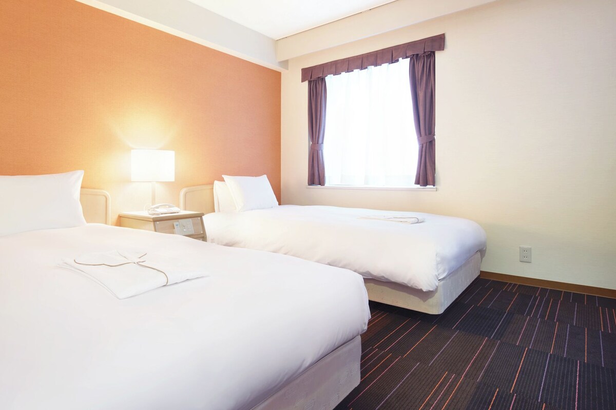 A hotel room with two beds, a bedside table with a lamp, and a window with curtains.