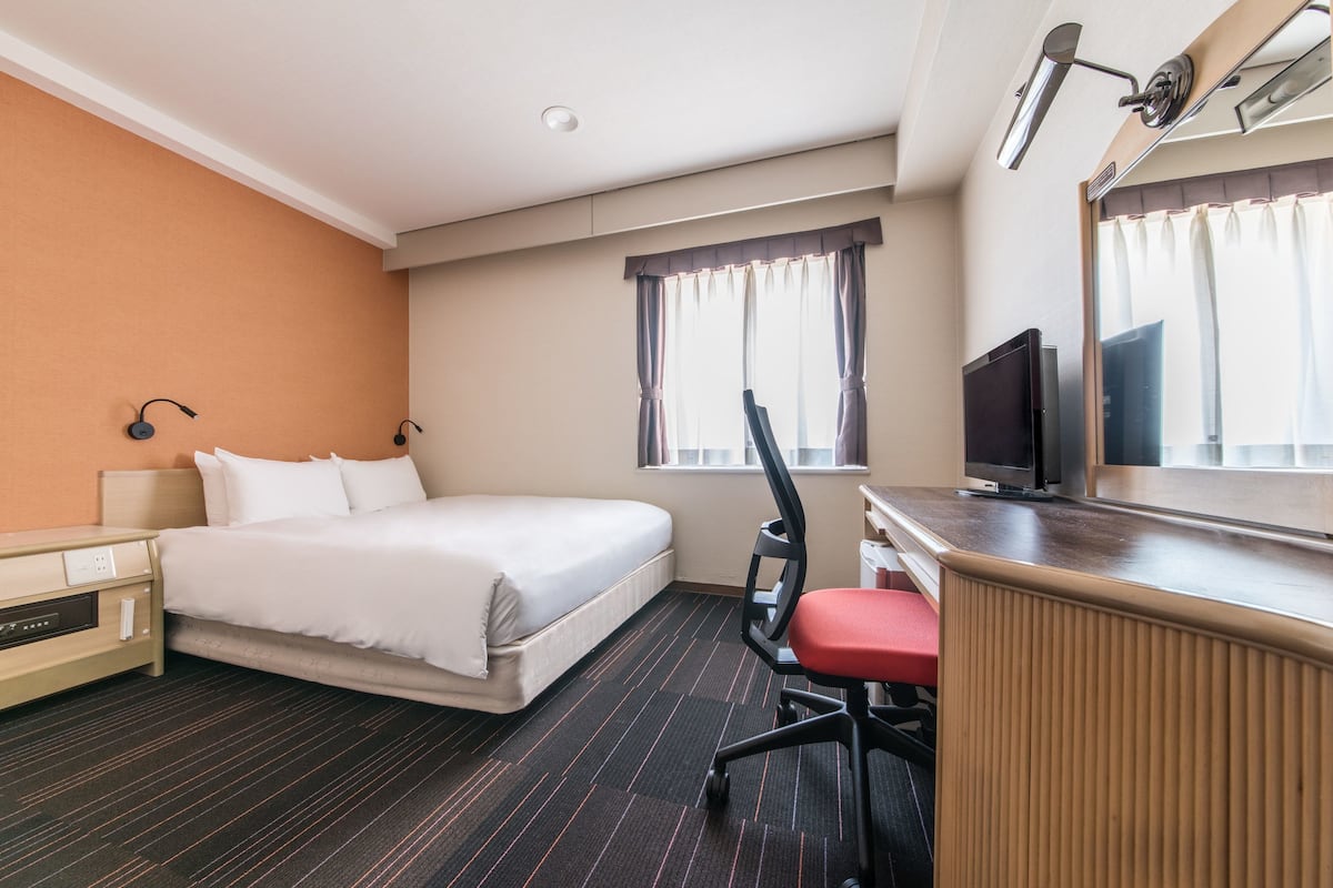 Una habitación de hotel con cama, escritorio con silla, televisor y ventana con cortinas.