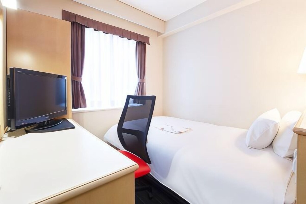 A hotel room with a bed, a desk with a computer, a chair, and a window with curtains.