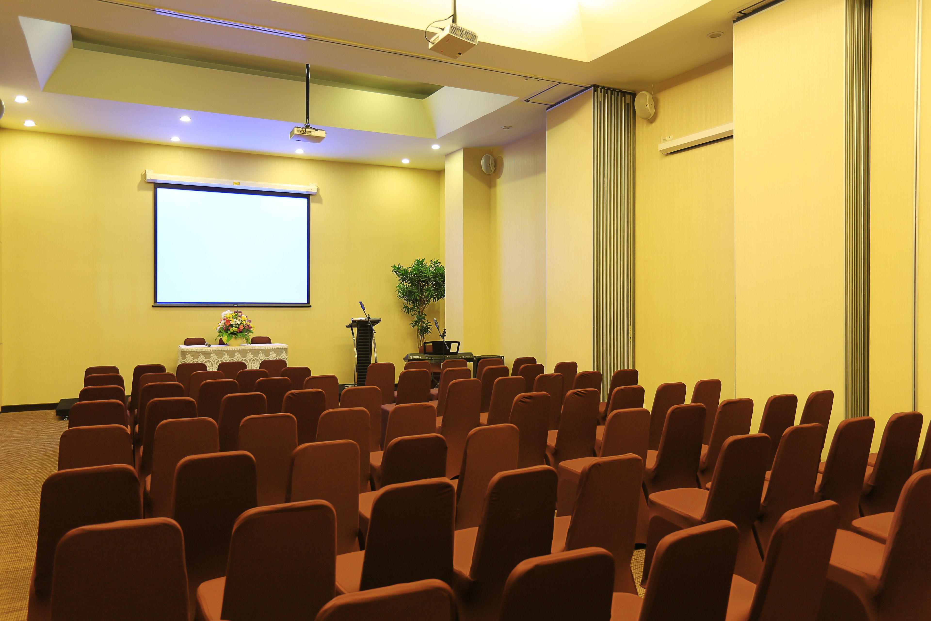 A conference room with rows of chairs, a projector screen, and a podium with a plant.