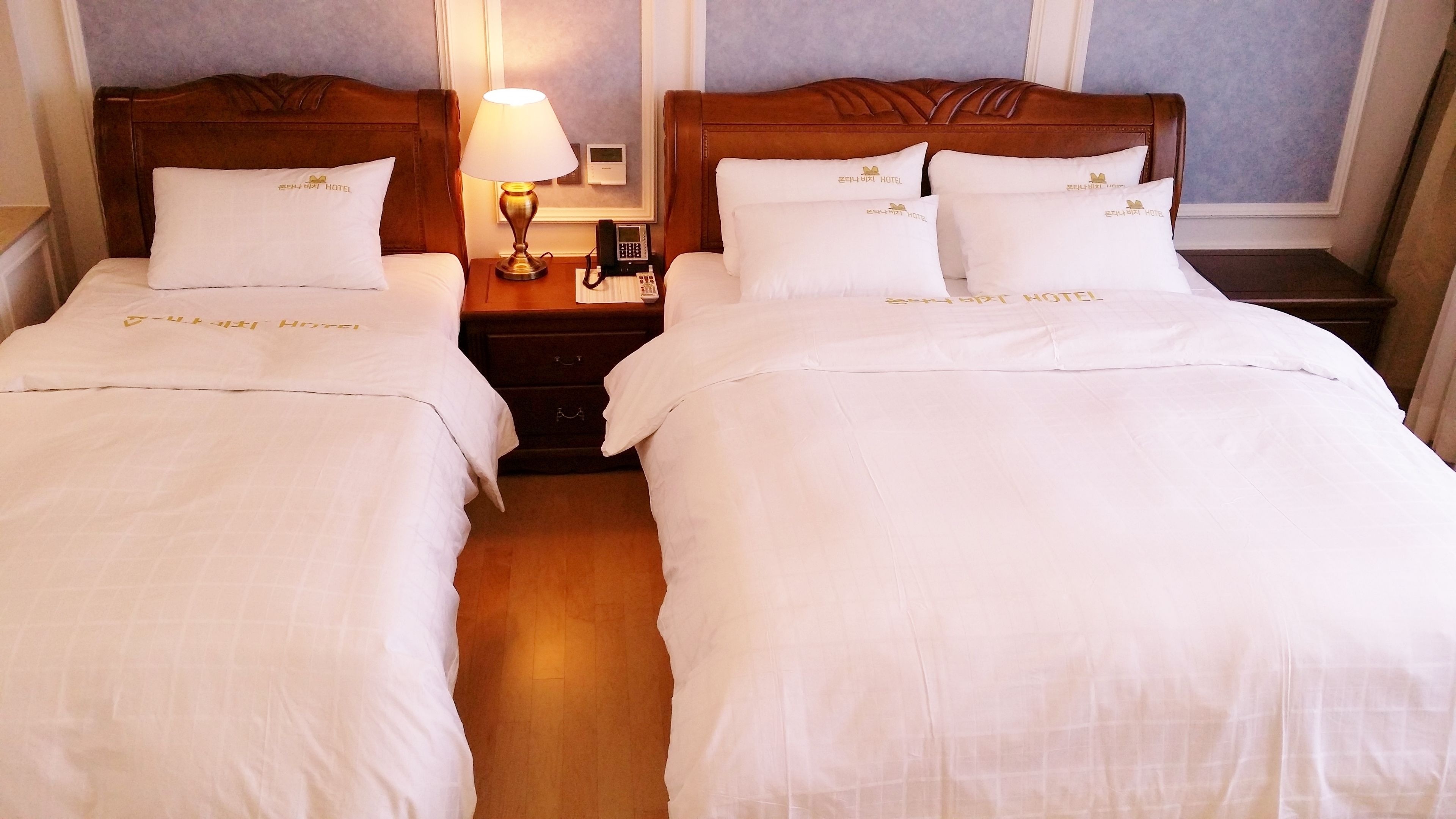 Habitación de hotel con dos camas, cada una con ropa de cama y almohadas que muestran la marca del hotel. Hay una mesita de noche con una lámpara y un teléfono entre las camas.