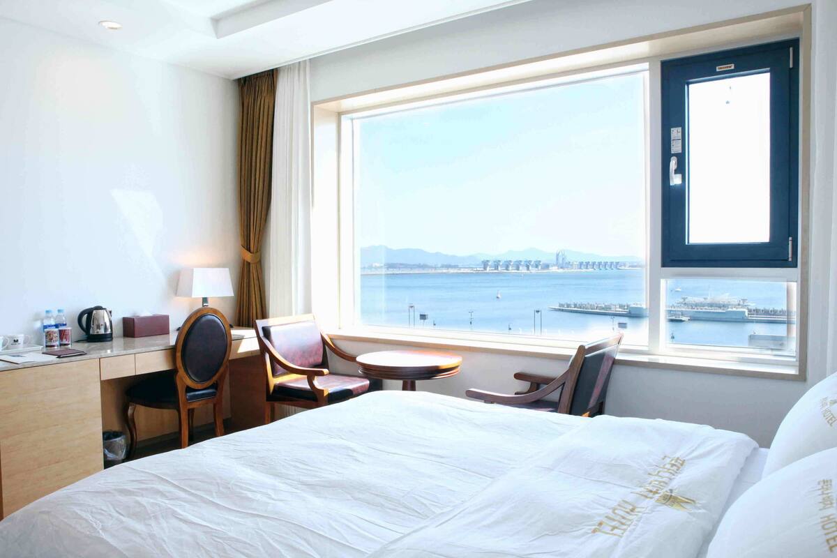 Ein Hotelzimmer mit Bett, Schreibtisch, Stuhl, kleinem Tisch und Blick auf ein Gewässer mit einer Brücke in der Ferne.