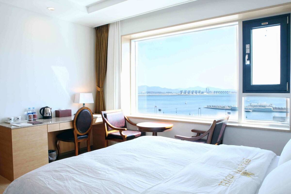 Ein Hotelzimmer mit Bett, Schreibtisch, Stühlen und einem großen Fenster mit Blick auf eine Brücke und Wasser.
