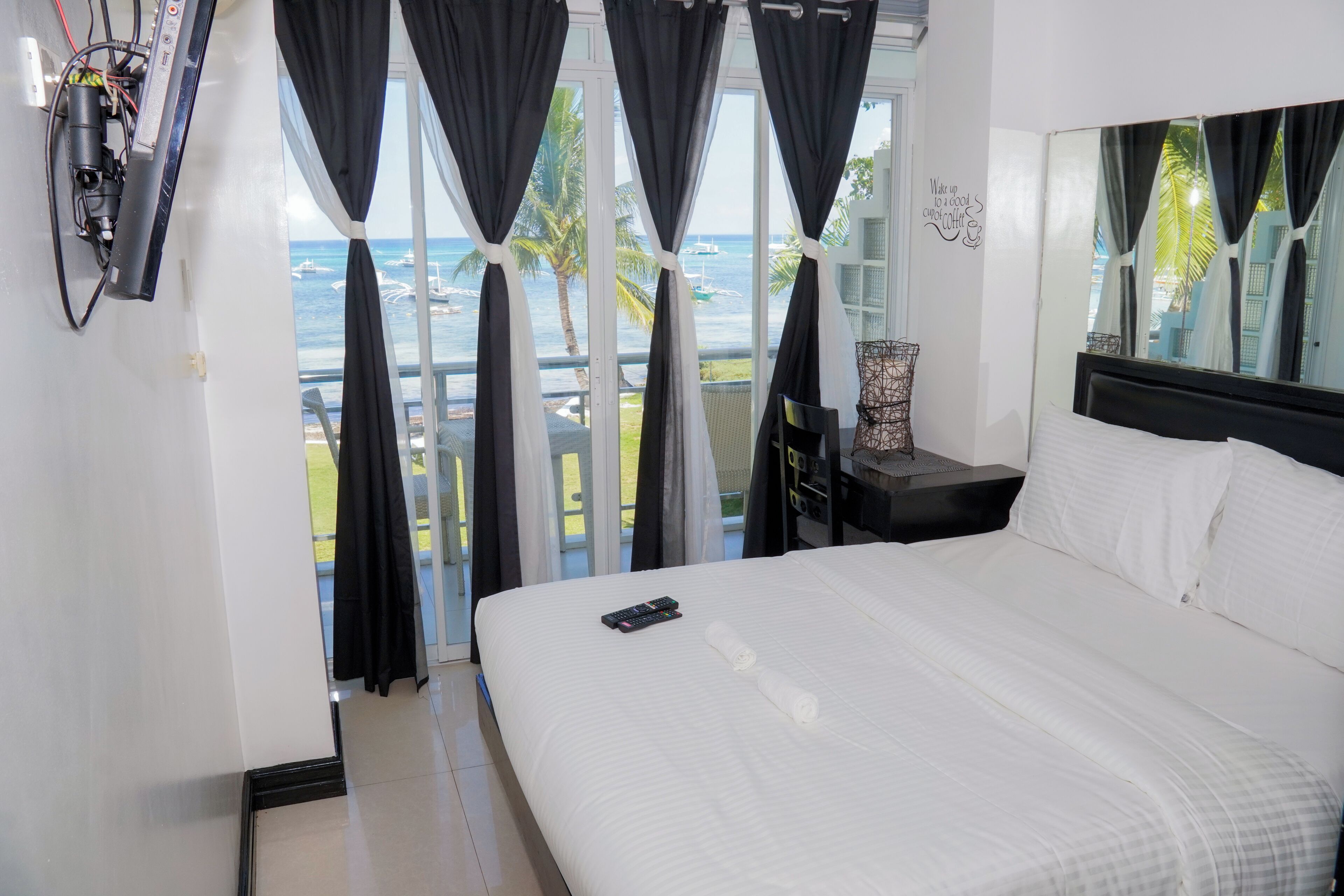 A hotel room with a bed, a nightstand, a lamp, and a view of the sea through large windows.